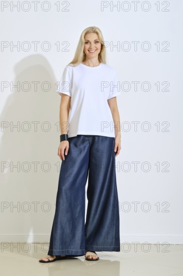 A smiling woman stands confidently in a bright studio space, wearing a simple white t-shirt and blue wide-leg pants. The soft natural light highlights her relaxed style and cheerful expression