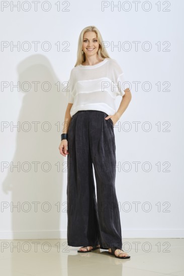A model stands confidently indoors wearing a light top and loose black pants. The setting features soft lighting and a plain background, creating a modern and elegant atmosphere