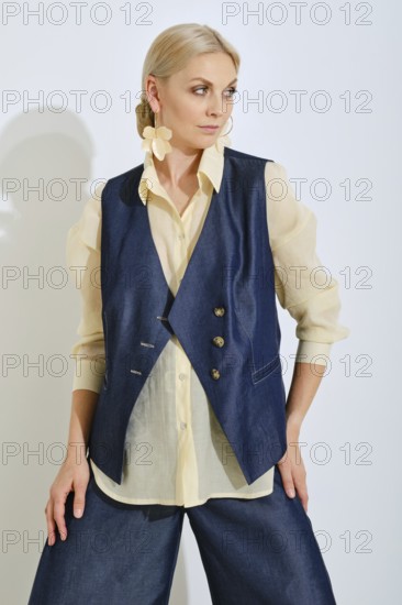 A fashion model poses confidently in a navy vest and light blouse. The background is bright and minimalist, emphasizing the trendy outfit in a casual yet fashionable way