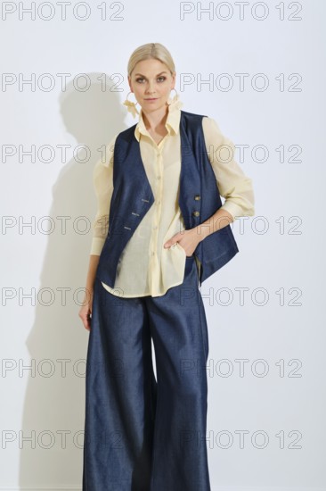 A confident woman showcases a chic fashion look featuring wide denim pants and a fitted vest over a light blouse. The studio setting has a bright background that highlights her outfit