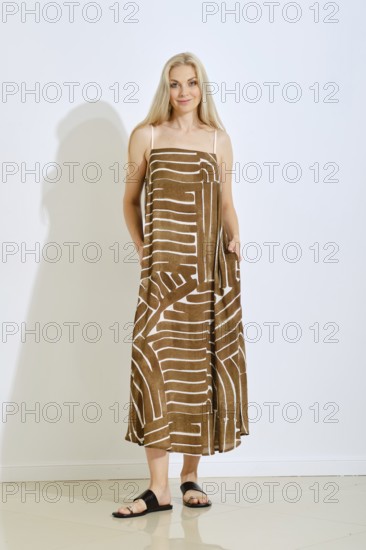A woman stands confidently in a brown sundress featuring a unique white pattern. The setting is a bright, modern studio with minimal decor, highlighting her fashion choice and relaxed pose