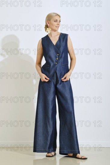 A woman stands gracefully in a stylish blue denim vest and wide-legged pants. She is indoors, showcasing her trendy outfit with a neutral background, highlighting her elegance and fashion sense