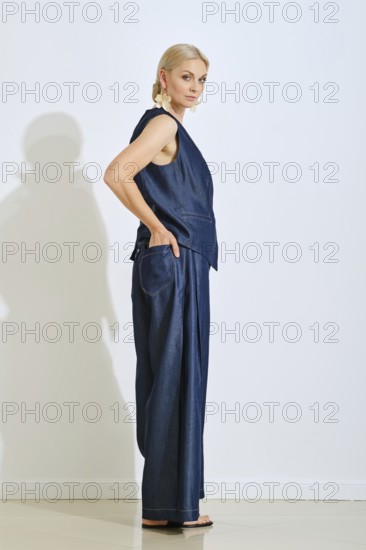 A model stands in profile in a modern studio wearing a sophisticated denim outfit. The light enhances the textures of the fabric as she poses with hands in pockets, exuding elegance and style
