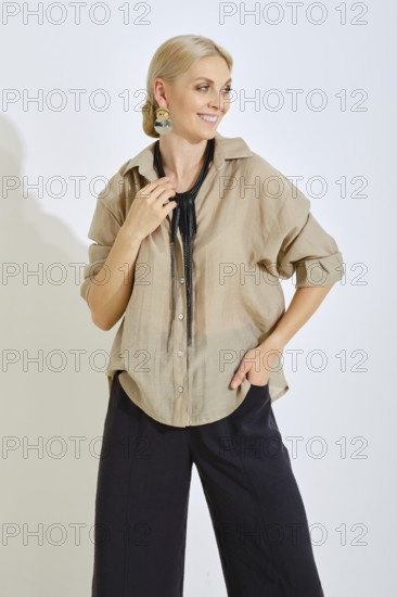 A woman is standing with a smile, dressed in a light beige shirt and dark pants. She wears a statement necklace and displays a relaxed yet fashionable look in a bright, minimalistic setting