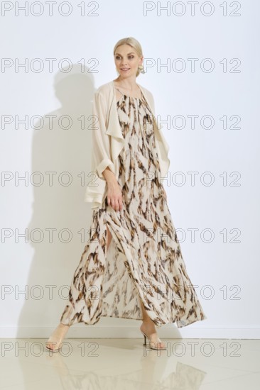 A stylish woman walks confidently in a beautiful, flowing dress with a unique pattern. The bright studio enhances her elegant look and the overall atmosphere is fresh and modern