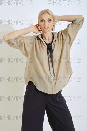 A woman strikes a confident pose while wearing a chic beige shirt complemented by bold accessories. The minimalist backdrop highlights her outfit, showcasing modern fashion aesthetics