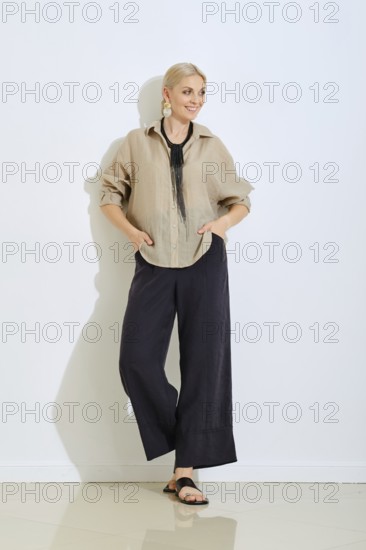 A woman stands confidently in a relaxed beige shirt and black pants, hands in pockets, on a clean white background. She accessorizes with a statement necklace, showcasing modern casual fashion