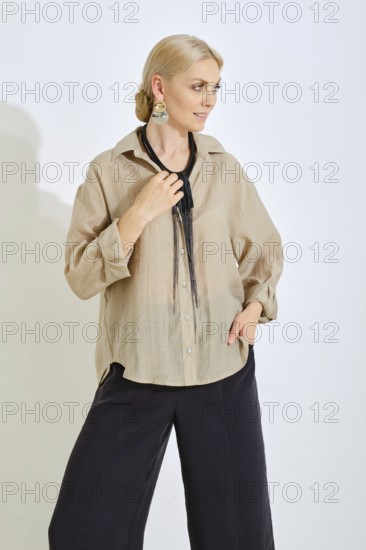 A woman stands gracefully in a modern studio, wearing a light-colored casual shirt and dark pants. She accessorizes with long earrings and holds a decorative necklace, exuding elegance