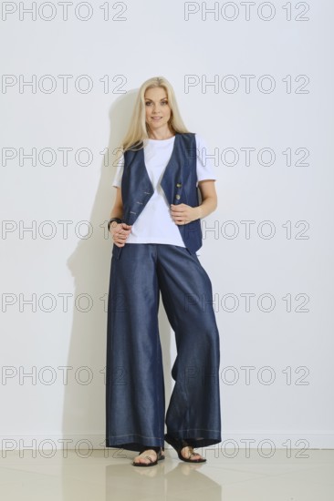 A woman stands confidently in a fashionable outfit consisting of a navy vest over a white shirt and wide-leg pants. The setting has bright lighting, enhancing her stylish look