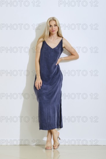 A woman stands gracefully against a plain white wall, wearing a long, sleeveless dress in a deep blue color. She has flowing hair and a confident posture as she poses simply, showcasing her outfit