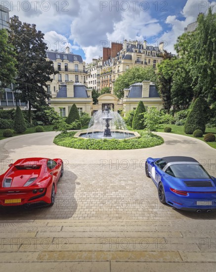 Two luxury sports cars parked in the driveway of an opulent Parisian mansion with view to a beautiful garden with classical fountain. Traditional Haussmann-style apartment buildings in the background, Paris, France