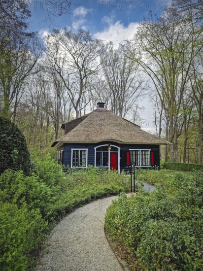 A winding paved path slopes down through the forest leading towards a rustic house nestled in the woods. Peaceful scene with secluded atmosphere, Lisse, Holland, Netherlands