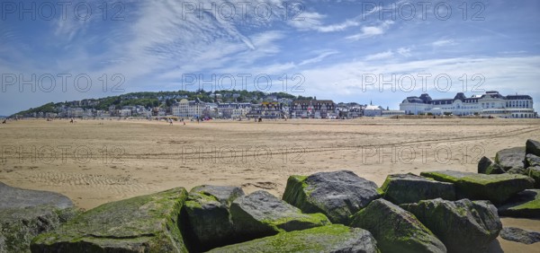 Panoramic view of Trouville-sur-Mer sandy beach, Normandy, France. Large, moss-covered breakwater rocks in foreground and charming town with traditional buildings in the background, Trouville-sur-Mer, France