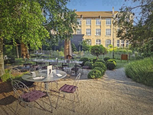 Rustic outdoor restaurant terrace with a classic, european garden aesthetic. The seating area features round bistro tables set on a gravel path. String lights overhead for a warm, inviting ambiance, Le Havre, France
