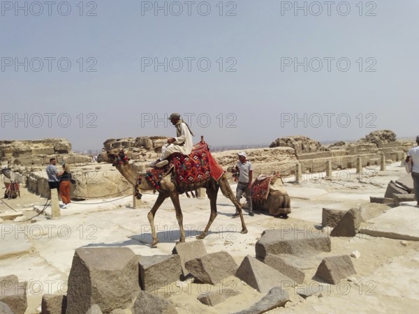 A group of Bedouins actively seek out tourists to ride their camels for a fee near the Great Pyramids of Giza, in the sands of the Sahara Desert. Nearby, the granite casing of one of the pyramids lies scattered, while tourists and travelers take selfies overlooking the archaeological site and the remains of Ancient Egypt. Cairo, Egypt