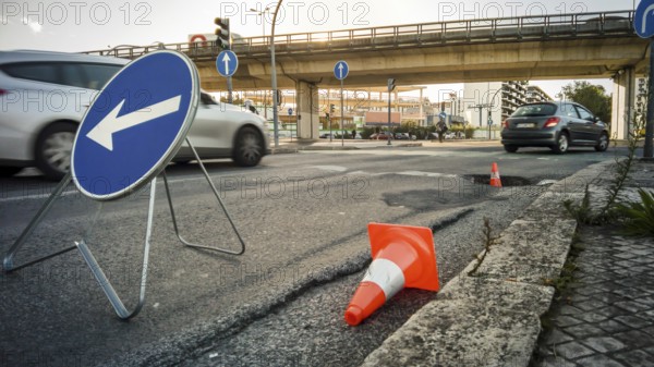 City street with large pothole marked by traffic cones and detour sign, cars passing by and overpass in background. Urban infrastructure issues, road safety, busy traffic hours, street repairs, traffic disruption, heatwave, asphalt melting, summer, Lisbon, Portugal