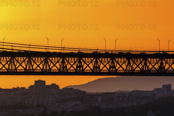 Bridge spans city skyline, silhouetted against vivid orange sunset. Soft city outlines and distant hills frame flowing late evening traffic, dynamic urban energy, tranquil evening light for unique harmony of architecture and nature. Urban landscape, Lisbon, Portugal