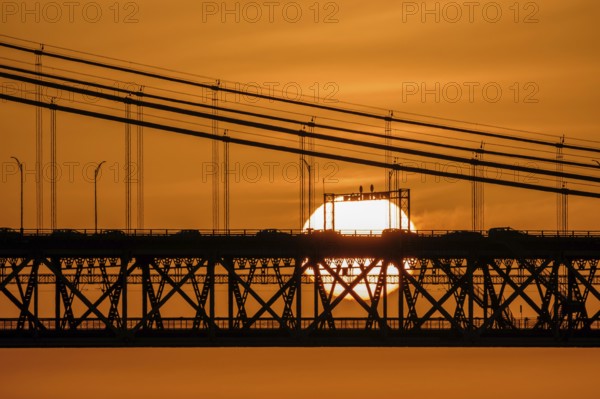 Glowing sun setting behind 25 de Abril bridge, Lisbon, Portugal, suspension cables, cars and bridge structure in silhouette. Bright orange sky, dramatic urban sunset over Tagus River. Close view, vehicles cross bridge, summer heat haze, bright sun