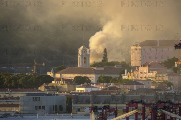 Lisbon church tower in sundown light surrounded by historic buildings and column of rising smoke in air. Soft sunlight, city architecture, beautiful tranquil urban skyline, risk of fires, damage, summer heatwave, emergency, firefighters alert