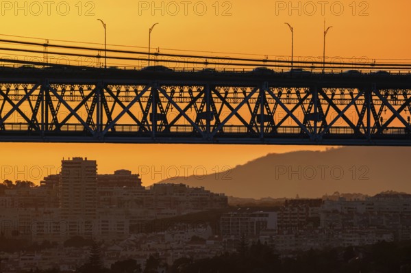 Striking close-up of famous 25 de Abril bridge, its silhouette against bright orange sunset sky. Lisbon, Portugal, cityscape and distant hills in hazy summer evening, blurred vehicles cross bridge, modern buildings, industrial structure, urban life