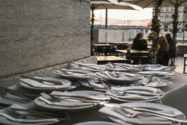 Plates neatly arranged with forks, knives, and napkins on stone countertop at outdoor restaurant in selective focus. Blurred background with European city square at sunset. Relaxed dining, people seated, umbrellas, travel destination, summer vacation, Lisbon, Portugal