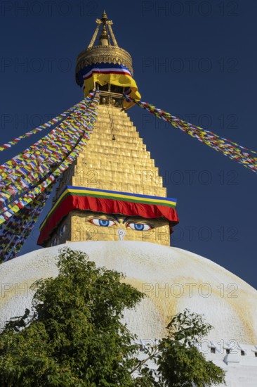 Boudhanath, the Boudha Stupa, the largest stupa in Nepal, Buddhist religious complex, UNESCO World Heritage Site and one of the main Kathmandu Valley tourist attractions. The upper part of the dome and the gilded, golden spire with Buddhist prayer flags stretched from the top. Eyes on the cubic part, the harmika, represent all-seeing, wisdom eyes of the Buddha. Seen in the afternoon on a clear, blue-sky, early autumn day. Kathmandu, Bagmati Province, Nepal