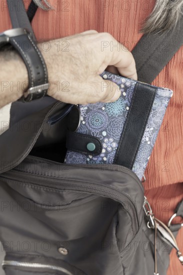 Pickpocket thief Person's hand storing a distinctive floral pattern wallet in a dark-colored shoulder bag, a watch visible on the wrist