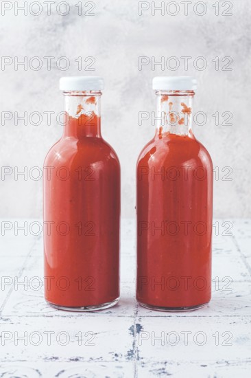 Two clear glass bottles hold vibrant red sauce, likely a spicy or tangy condiment, in a well-lit kitchen area. The smooth surface enhances the color and freshness of the sauce