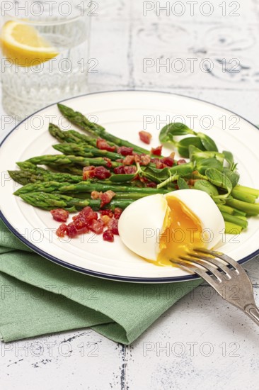 Poached egg with fork, asparagus, bacon slices and herbs, on a white plate, breakfast, close-up, no people
