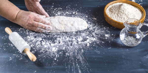 Female hands preparing fresh artisanal bread dough on a dark kitchen table. Baking ingredients flour, water, and a rolling pin are present