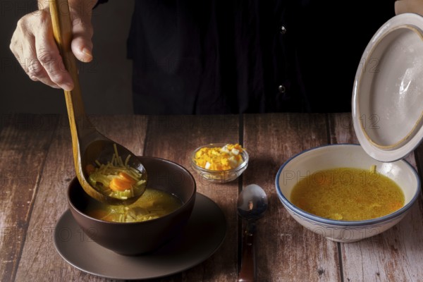 Woman serving chicken soup with a wooden spoon in a brown bowl on a wooden table