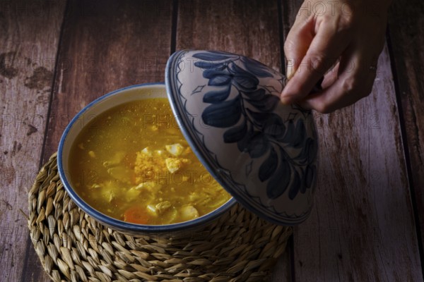 Woman opening the lid of a tureen with chicken noodle soup and vegetables on a wooden table