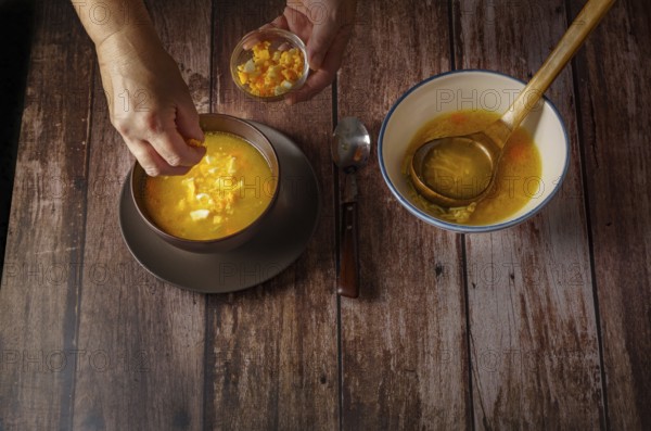Woman pouring hard-boiled egg into a chicken noodle soup in a brown bowl on a wooden table and a white soup tureen