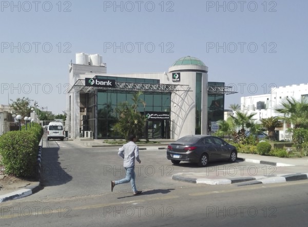 A local Egyptian bank, specializing in housing development and owned by major foreign investors Alpha Oryx, is surrounded by date palms and near a busy highway. A lone man runs down an alleyway toward the bank. It is located in the Hadaba district of the Egyptian tropical resort town of Sharm el-Sheikh. South Sinai. Egypt