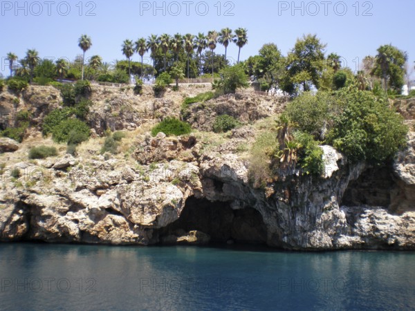 A panoramic view of a grotto with caves in the mountain cliffs jutting out from the azure and turquoise waters of the eastern Mediterranean. Palm trees grow on the cliff top. A breathtaking landscape that draws tourists and travelers from around the world to see this natural landmark of the Turkish Riviera. Antalya Province, Türkiye