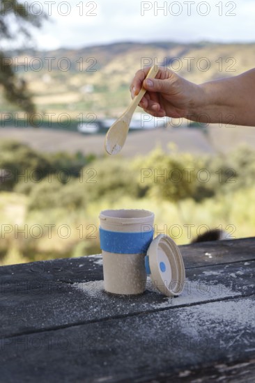 Person's hand adding a white powder with a wooden spoon into a reusable eco-friendly cup, enjoying a picnic in nature