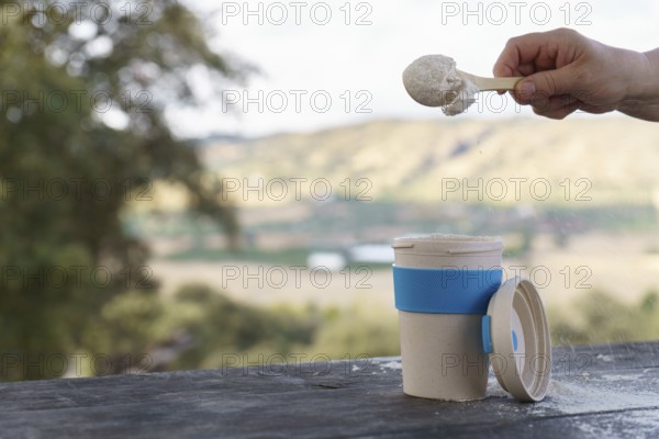 Hand scooping protein powder into a reusable cup on a wooden table, preparing a healthy lifestyle drink with a scenic background