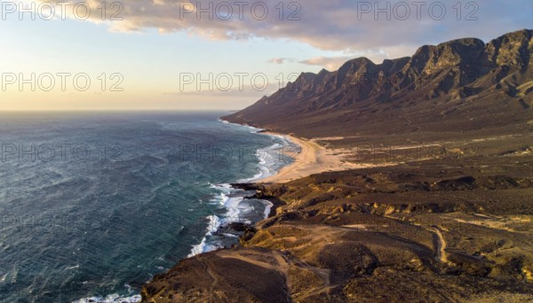 Desert landscape converges with ocean along a rugged coastal area, shore break at cliffs, blue cloudy sky at sunset, dry landscape in an aerial drone view, AI generated