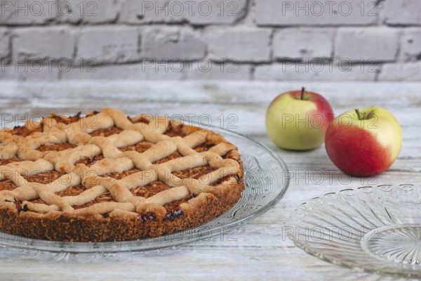 Apple cake on a glass plate with two apples next to it in front of a rustic brick wall