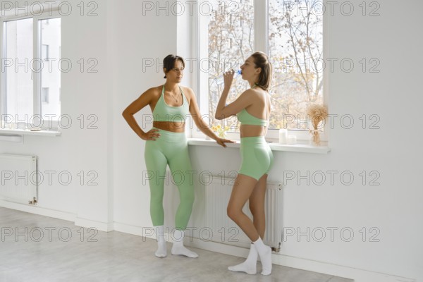 Two women in green activewear take a break during their fitness stretching class. They are standing near a window, enjoying a moment of rest and hydration in a bright studio environment