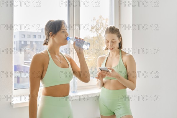 Two women take a break during a fitness stretching class in a bright indoor studio. One drinks water while the other checks her phone, enjoying the pause in their workout session