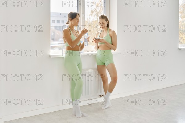 Two women in matching green workout outfits take a break during a fitness stretching class in a bright studio. They talk and hydrate by the window, enjoying time together after their workout session