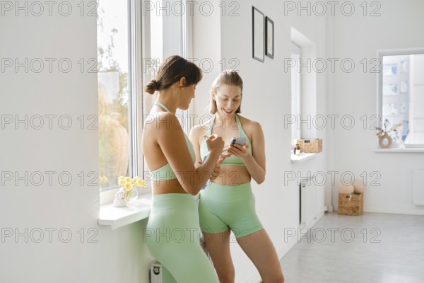 Two women in matching green workout outfits take a break during a fitness stretching class. They are smiling and looking at a phone while relaxing by a large window with natural light