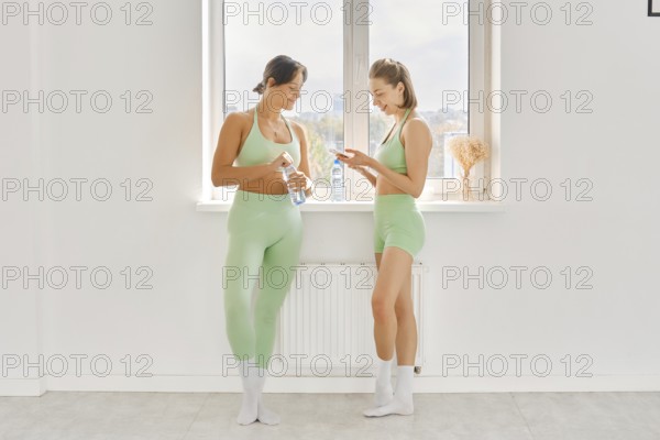Two women pause during their fitness stretching class in a bright and spacious studio. They are dressed in matching light green workout outfits and relax by chatting and checking their phones