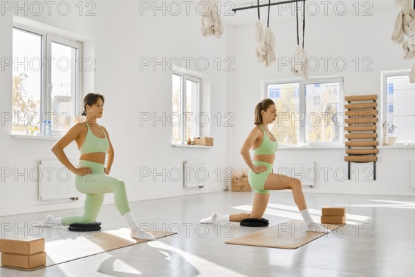 Two women practice lunges in a bright fitness studio. They use yoga mats and blocks for support. The room is spacious with large windows, creating a calm and inviting atmosphere