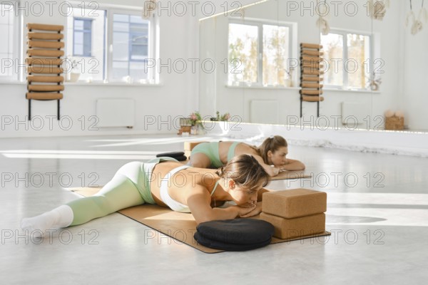 In a spacious and well-lit yoga studio, two women are engaged in a stretching routine on mats. They focus on their practice, surrounded by peaceful decor