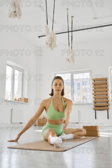 A young woman is seated on a yoga mat in a spacious, well-lit studio. She is stretching her body with one hand supporting her and the other reaching out, focusing on her breathing