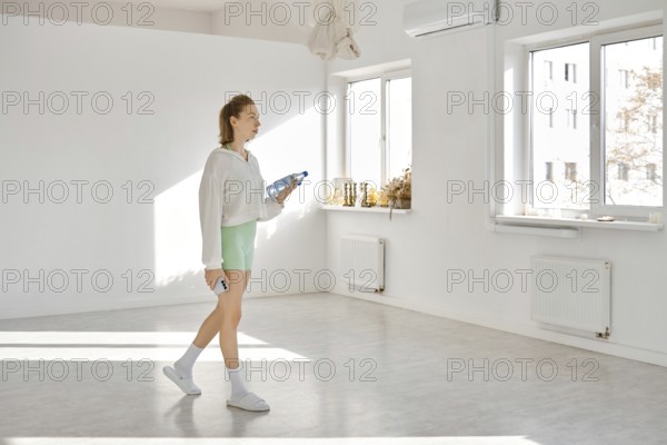 In a bright studio with white walls and sunlight streaming through large windows, a young woman walks with drinking water bottles