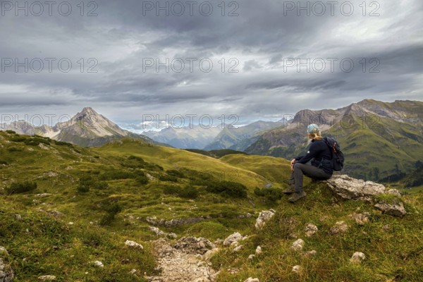 A hiker sits on a hill with a view of a mountainous landscape under a grey cloudy sky, Schröcken, Austria