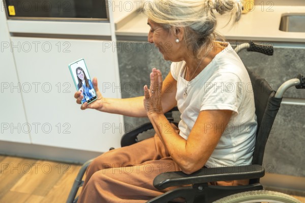Senior woman in a wheelchair laughing and waving during a video call on her smartphone, connecting with a doctor or healthcare provider for remote assistance and virtual medical consultation